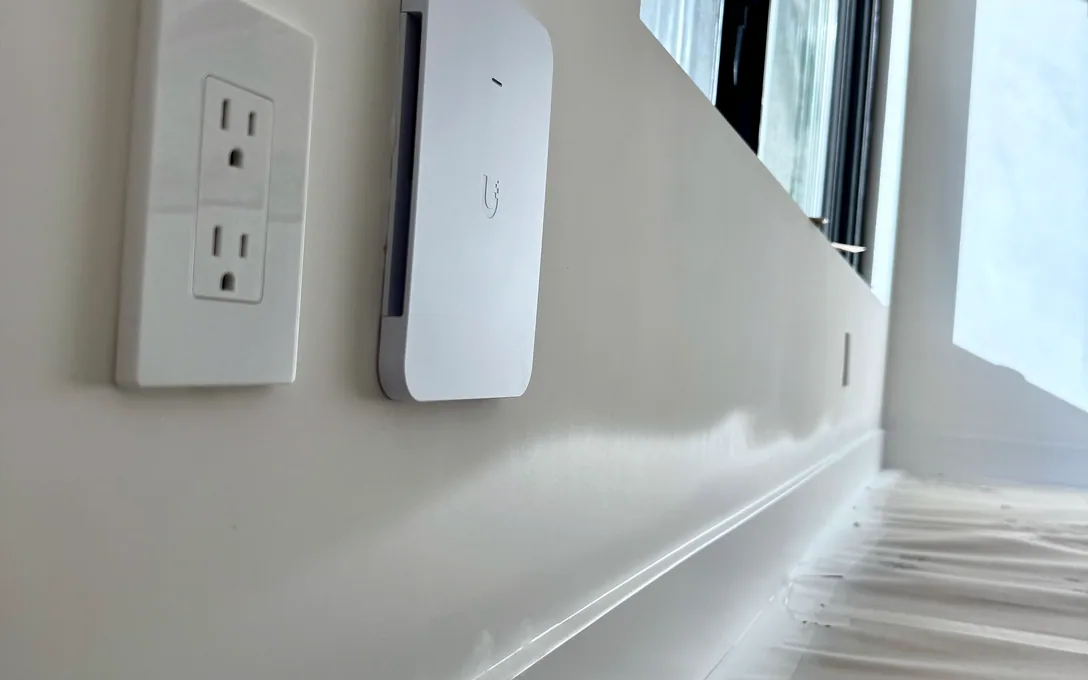 White UniFi wall access point mounted beside a double outlet on a white wall, floor covered in protective paper.