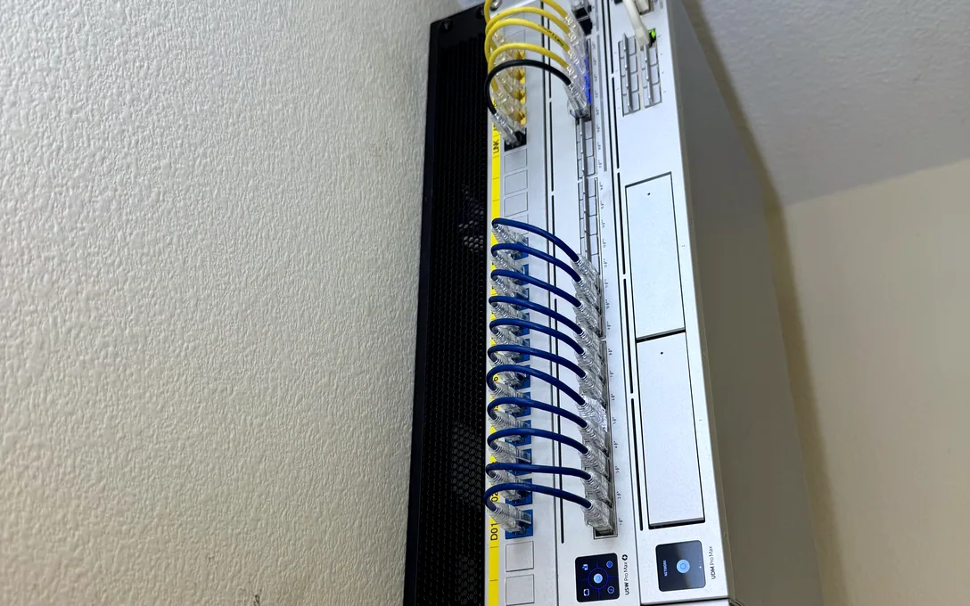 Black patch panel on a white wall with blue Ethernet cables routed through a side cable manager.