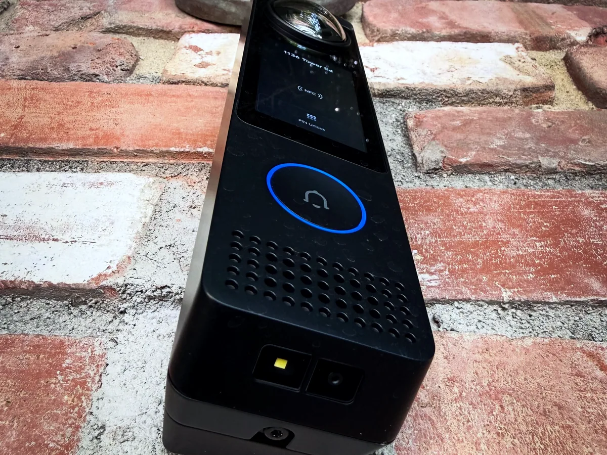 Black UniFi Access intercom reader with a blue ring button mounted on a red brick wall.